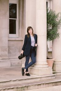 Lucy perched against wall holding camera, owner of Lucylou Photography - Hampshire Wedding Photographer